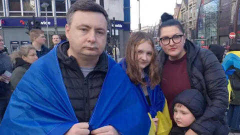 A man, woman, a girl and boy stand together. The man and girl have the Ukranian flag draped over their shoulders. People are standing around them.