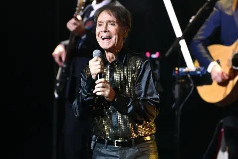 Getty Images Sir Cliff Richard on stage at a concert holding a microphone and smiling. He wears a black jacket with gold sequins and dark shiny trousers. Two guitarists are behind him.