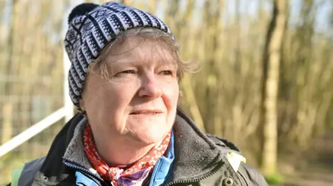 Diane Taylor is wearing a black and white bobble hat with a thick coat on top of a fleece. She is stood with woods behind her.