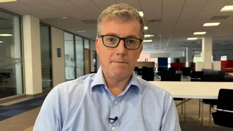 Dr Chris Williams is looking at the camera and not smiling. He has short grey hair. He is wearing black square frame glasses and a light blue shirt.