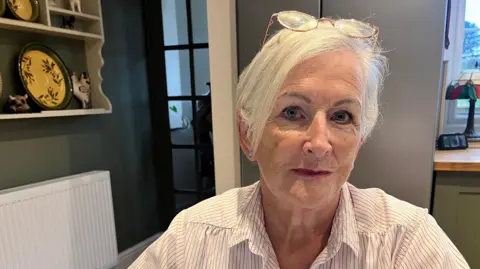 Julie Doorne sits in the kitchen at her Lincolnshire home. She has short, white hair and wears a white and grey striped blouse and pink-rimmed glasses on the top of her head.