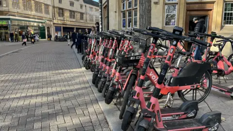 Tom Jackson/BBC A group of pink e-scooters and e-bikes are parked on a stretch of pedestrianised city street. Several people are walking in the area.