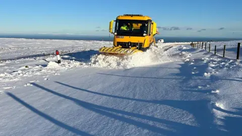 A yellow snowplough pushing through a thick blanket on snow on the Mountain Road. There are farmers' fenceposts at the side of the road.