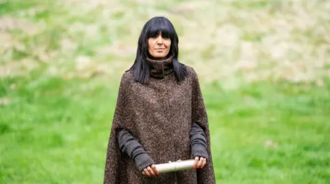 BBC/Studio Lambert Claudia Winkleman, a woman with shoulder length black hair and a fringe which covers her eyebrows. She is wearing a brown woollen cape and holding a scroll in both of her hands. She is stood on grass.