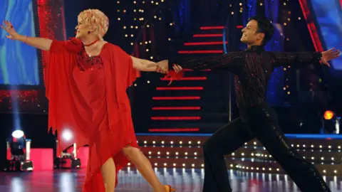 Gloria Hunniford and Darren Bennett perform during 'Strictly Come Dancing'. Gloria is wearing a red dress and Darren is wearing a black shirt and black trousers. They are holding hands with the other arms in the air