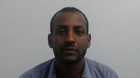 Amaniel Tewelde's custody mugshot. He's wearing a blue shirt with a pattern on the inside. He has a dark goatee style beard and short black hair.