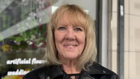 Carole Burke has a blonde bobbed haircut and wears a black leather jacket.