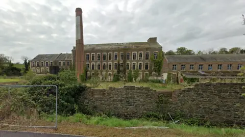 Google Maps The former Sinton's Mill pictured in April 2022. The complex is a mixture of brick Victorian buildlings of different heights surrounding a red brick chimney stack. Many of the windows are broken and there is an uneven wall surrounding the site.