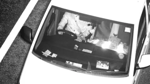 A person sat at the wheel of a car. They are wearing a seatbelt and have one hand on the wheel, while the other hand appears to be holding a mobile phone. There are multiple items on the dashboard and in the passenger seat.