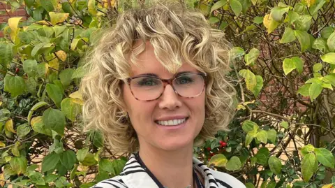 A picture of a blonde woman, with a short curly blonde pixie hair cut. She is wearing a white blouse with black stripes and has glasses on.