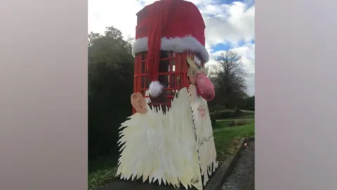 Cary Otis A Santa face on a telephone box. There is a red hat on the top of the box and the Santa model has large eyes, a big red nose, ears, and a beard
