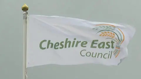 BBC A white flag with Cheshire East Council's name and logo on it in green and yellow.