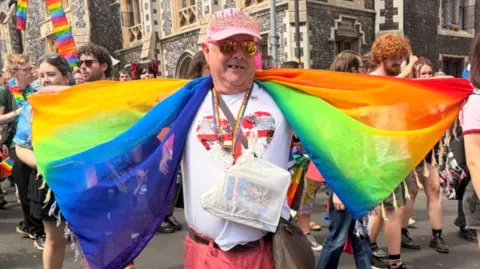 Harriet Morter/BBC A man in the middle of the march at Norwich Pride. He is looking directly at the camera and is wearing a white T-shirt with a red heart in the middle, a pair of red trousers, a pink baseball cap with a crown on it and draped over his arms and back is a rainbow flag.