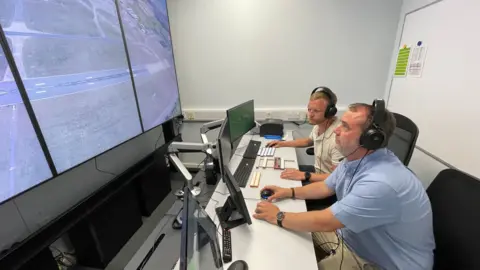 Two members of staff are sitting with headsets on looking at a big screen which has the airport runway showing. They are in the air traffic control simulator room.