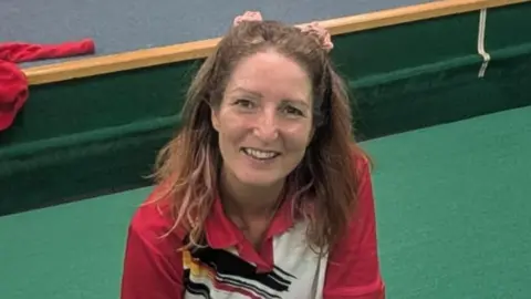 Submitted Emma Price kneeling on green felt at the indoor bowling club. She is wearing a red sports shirt and smiling up at the camera. He brown hair is tied back with two pink scrunchies. 