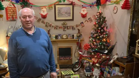 George Keys in a blue sweater standing behind the 1960s living room display he made from various items. 