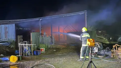 Roger Apsey A firefighter is putting out a fire at a barn at night. The firefighter is wearing a black and yellow uniform and a helmet. The barn is burning with orange flames and thick smoke visible.