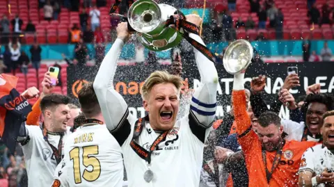 Byron Webster holds the National League promotion final winners' trophy aloft cheering, while teammates around him celebrate 