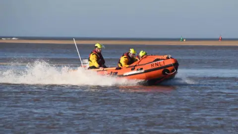 Three people onboard a small orange lifeboat which is travelling at speed across the water.