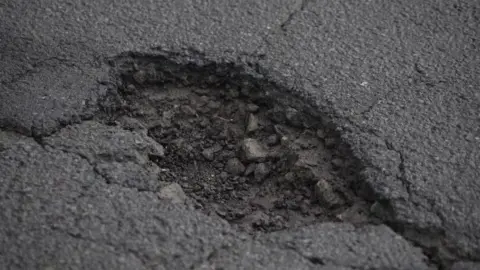 A close‑up view of a pothole in a road surface. It shows cracked edges and exposed loose stones within the damaged tarmac.