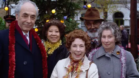 BBC Three women and a man stood in a row smiling looking ahead. They are wearing jackets with coloured tinsel round their necks. They are stood outside and in the background is a tree with coloured lights on. 
