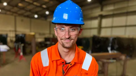 Martin Giles/BBC Callum Payne stands in an engineering workshop wearing a blue hard hat and luminous orange overalls. He has protective glasses over his eyes. A small goatee beard is visible on his face.