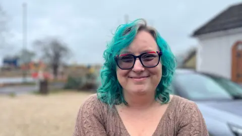 George Carden/BBC Heather Pencil standing in her drive, she has a brown top on, glasses and turquoise coloured shoulder length hair 