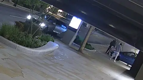 A car can be seen smashed into the front of a shop, with men surrounding it. The image is taken from CCTV footage which is taken on a slant.