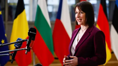 Getty Images Rachel Reeves, UK chancellor of the exchequer, stands in front of a podium with several microphones for media outlets. She wears a burgundy suit and white blouse and her fringe is swept to the side. 