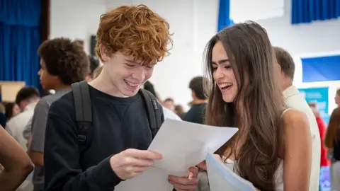 A young women and a young man in a crowded gymnasium looking down at a sheet of white paper the boy is holding. they both have broad smiles