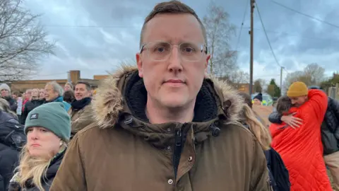 Shaun Whitmore/BBC Scott Lyons is wearing a winter coat with fur trim on the hood and glasses. He is looking solemnly at the camera, while protesters are in the background.