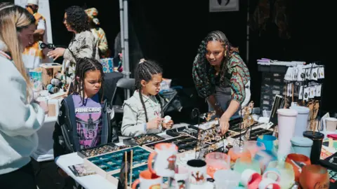 We Invented The Weekend Two young girls stand looking at rows of jewellery on a table. Two adult women stand next to them, and one of the women is pointing at an item of jewellery as if to show the girls.