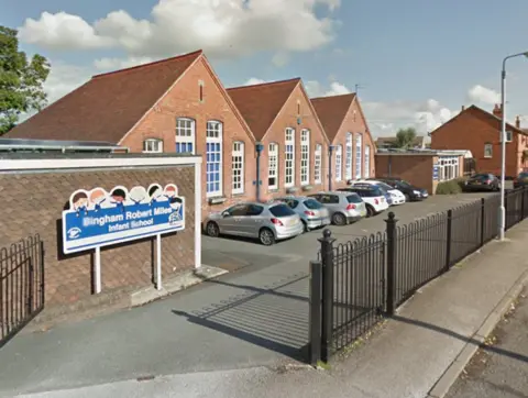 Google A school building, with a school sign outside, with cut-out children over the school's wording in white on a blue background. The school is one building in red brick and tall, white-framed windows and three separate arched rooftops. The building is by a grey tarmac car park, where six cars are parking in a row. 