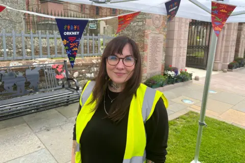 A woman in a high-viz vest with glasses on and blue and red bunting hanging from a marquee in the background.