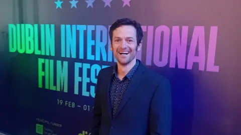 Mark Lowery A man at a film premiere who is standing in front of a promotional board that reads: Dublin International Film Festival. He has short, dark hair and wears a navy suit with a blue shirt underneath. 