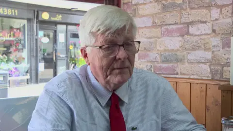 An older man in a light blue shirt and a red tie is sitting at a table in a cafe being interviewed. There are shop fronts visible through the window behind him. 