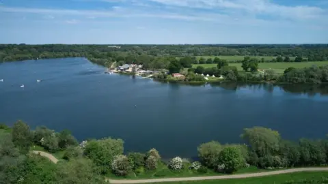 NENE PARK TRUST A wide expanse of lake can be seen from a bird's eye view, with trees, fields and a small number of buildings on either side. 