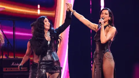 Getty Images Two women, Cher and Dua Lipa. Cher has long black wavy hair and wears a short leather skirt with a buckle. She holds the hand of the second woman, Dua Lipa. She is wearing a black sparkly swimsuit. 