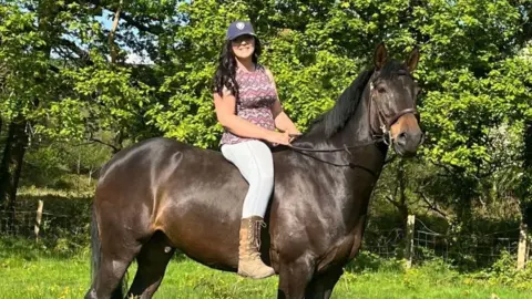 Facebook A woman, Tasha Lee Hopkins, sits without a saddle on a large, dark brown horse in a field and smiles into the camera. She is wearing a cap, patterned top, light blue leggings and tan lace-up boots. Her long brown hair is down.