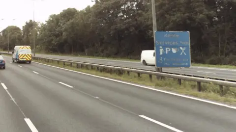 National Highways A picture of the M6, with vehicles travelling along it. A blue sign is located on the central reservation.