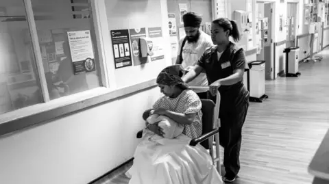 James Clifford Kent A midwife in scrubs pushes a mother in a wheelchair through a hospital corridor; the mother is cradling her newborn baby while the father walks closely behind them.