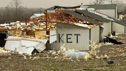 A building is in ruins after a tornado strike, its roof is ripped off and insulation is strewn about.