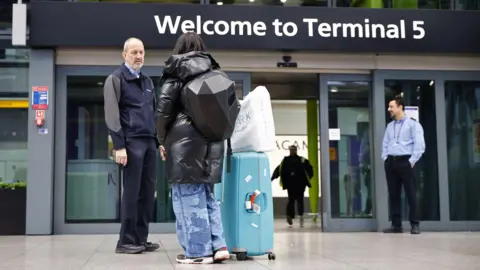 EPA Seorang penumpang dengan koper biru dan tas putih berdiri dan berbicara dengan anggota staf di luar pintu masuk ke Terminal 5 di Bandara Heathrow