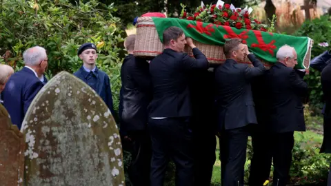 Media Wales Hefin David's coffin draped in a Welsh flag with red roses on the top, being carried by six people, with three men seen from the side. A young air cadet can be seen to the coffin's left.