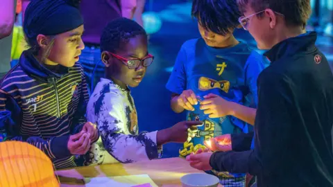 Four children gathers around a table, closely examining small objects illuminated by colorful, low light.