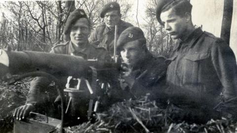 John aiming a machine gun with three other soldiers next to him