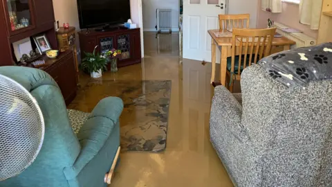 The inside of a lounge, filled with water, there is dirty water on the ground. There are two chairs, a table, chairs, a carpet and furniture. 