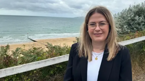 Millie Earl standing on the seafront. She has long fair hair and round glasses and is wearing a white top and black blazer. In the background is the beach and the sea.