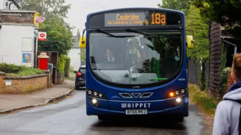 Cambridgeshire and Peterborough Combined Authority A blue single-decker bus with the destination Cambridge via Cambourne and Comberton written on a digital display above the windscreen. It is facing the camera on a narrow village road, with a post office and post box on the side of the road. 