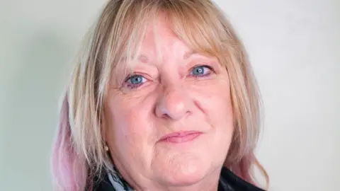 Hoskins Funerals A woman with blond hair and pink highlights, wearing a blue scarf, looks straight at the camera and smiles. 